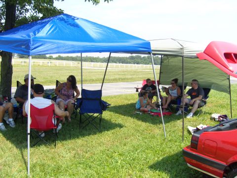 2007 Ford Mustang - Attendees relax under shade canopies at an outdoor event, with a red vehicle partially visible.