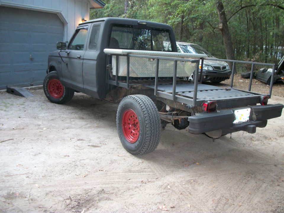Flatbed pickup truck with red wheels parked by a garage, another car behind it