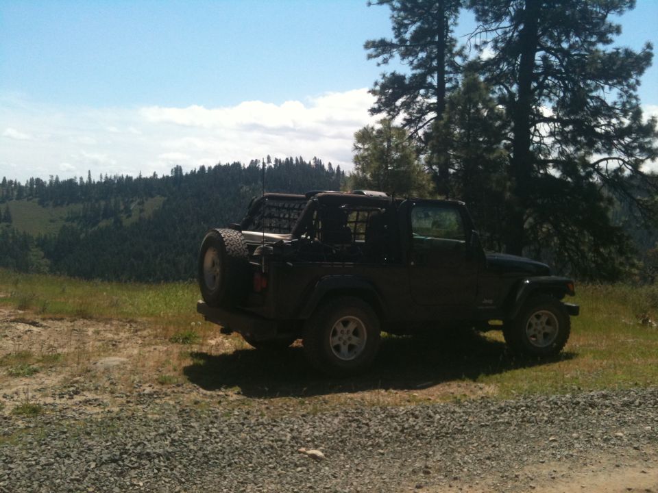 Black Jeep Wrangler parked on a gravelly area with a wooded hillside in the background