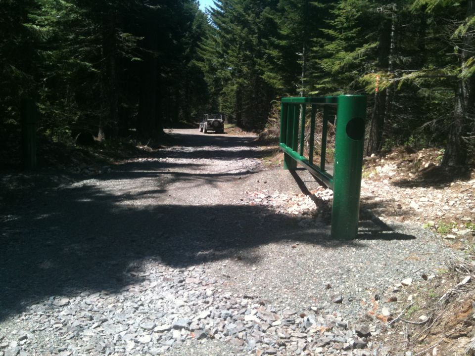 2005 Jeep Wrangler on a gravel road leading through a forest, with a green gate in the foreground