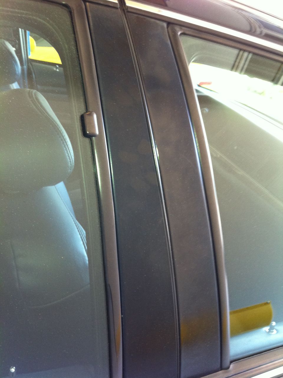 Dark colored car door pillar with visible smudges and a reflection of a leather seat. A yellow object is visible through the window