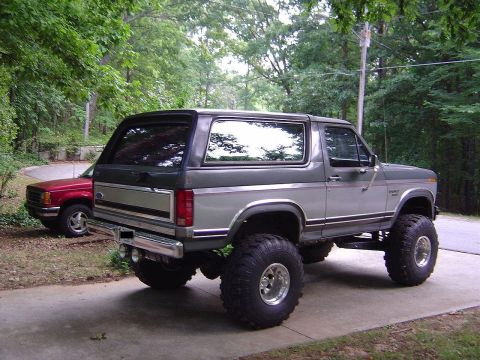 1986 Ford Bronco - A lifted 1986 Ford Bronco is parked on a driveway, with a red pickup truck visible behind it.