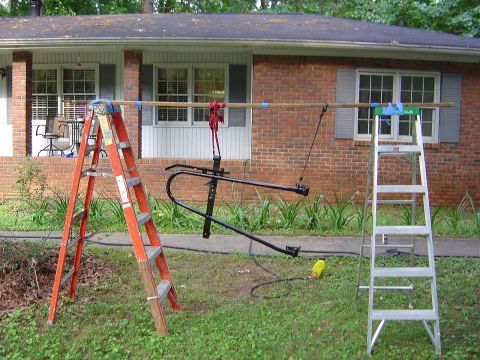 1986 Ford Bronco - A black spare tire carrier assembly is suspended by a rope and pulley system, with ladders and a house in the background.