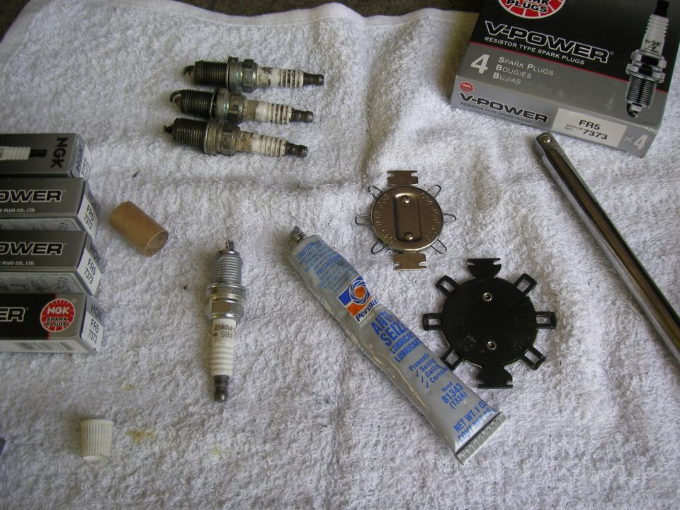 Four used spark plugs, one new spark plug, two boxes of NGK spark plugs, a tube of Permatex anti-seize lubricant, a spark plug gap tool, and a socket extension on a white towel