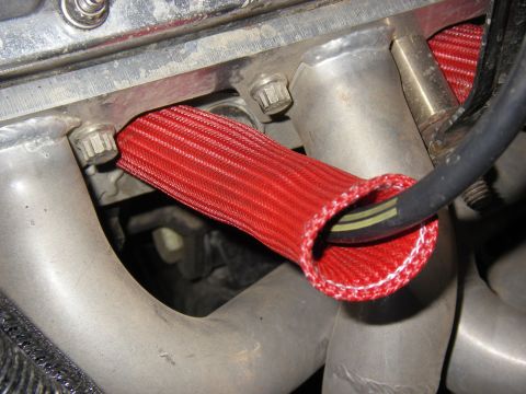 1990 Ford Bronco - Close-up of an engine spark plug wire protected by a red sleeve, near the exhaust manifold.