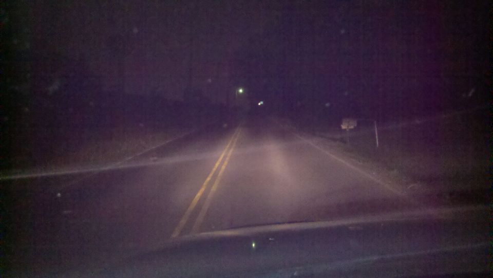 Night view over a vehicle's hood of a dark two-lane road lit by headlights, double yellow line ahead