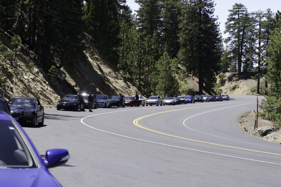 Blue car side mirror and hood in foreground, line of cars parked on winding mountain road with trees and rocky slope