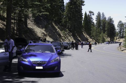 2006 Cadillac STS - A group of cars are parked along a winding mountain road, with trees and a hillside in the background.
