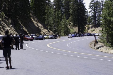 2006 Cadillac STS - A group of cars are lined up on a scenic mountain road, with onlookers gathered nearby.