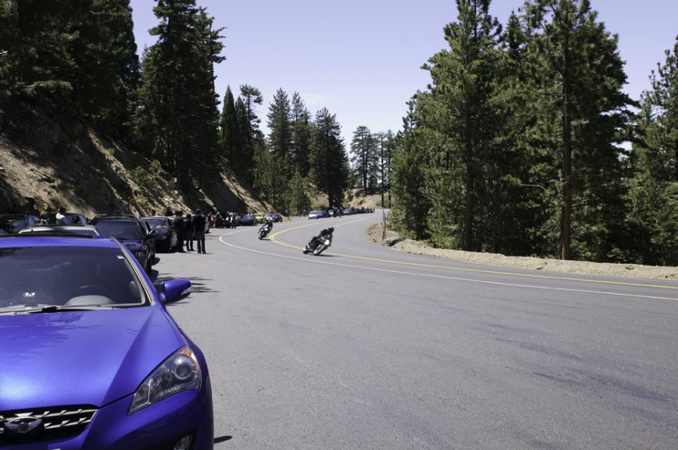 Motorcycles riding on a curved road in a forested mountain area. Cars are parked on the side