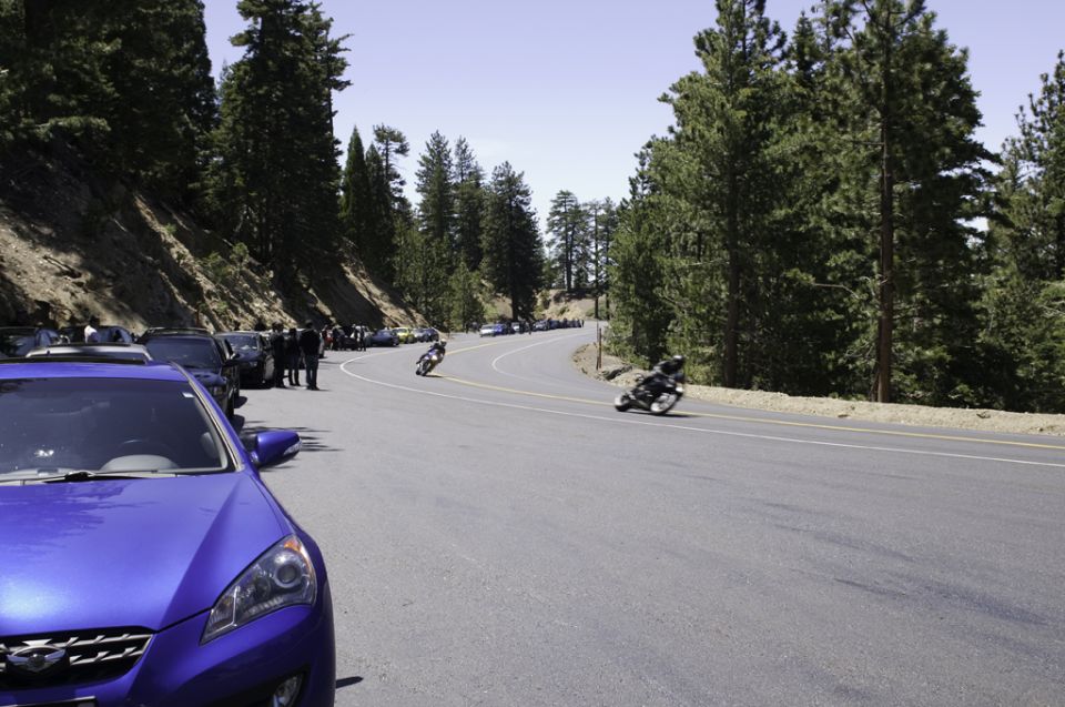 Blue car in foreground, motorcycles racing on a mountain road with pine trees