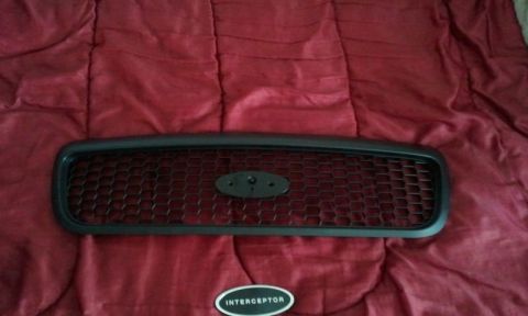 1998 Ford Crown Victoria - An unpainted black honeycomb-mesh Ford grille laid face-up on a red bedspread, with the bare oval badge mount in the center. Below it sits…