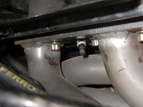1990 Ford Bronco - Close-up of a spark plug connected to an exhaust manifold, showing metal pipes and bolts.