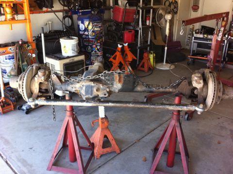 Suzuki Samurai - A complete used front solid axle assembly resting on red and orange jack stands in a garage, still greasy with knuckles, disc brake rotors,…