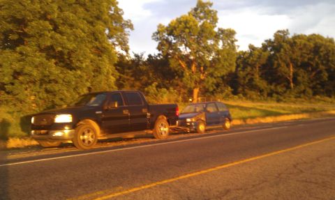 1991 Ford Festiva - A black pickup truck tows a small blue hatchback along a rural road during golden hour.