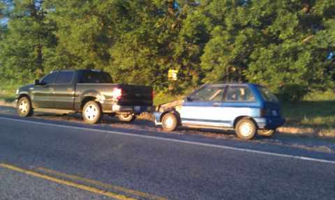 1991 Ford Festiva - A blue 1991 Ford Festiva is being towed by a dark-colored pickup truck on the side of a road.