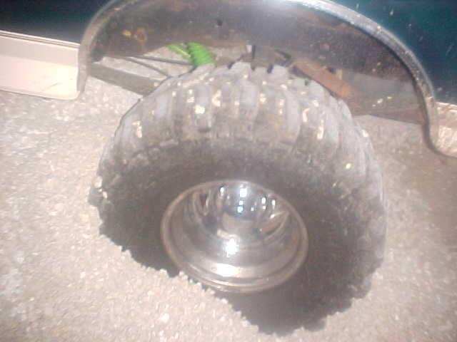 close-up of a large, knobby off-road tire on a chrome rim, with part of a dark vehicle body visible above