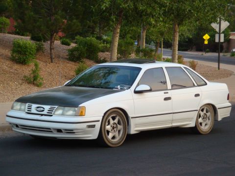 1990 Ford Taurus - This white sedan has a contrasting black hood and aftermarket wheels, suggesting some customization.