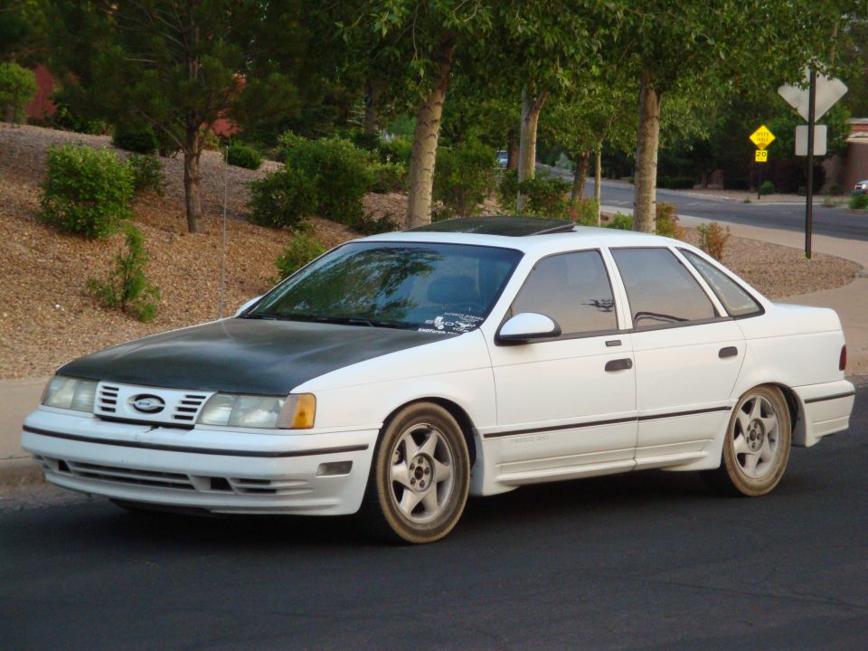 White Ford Taurus with a black hood and aftermarket wheels parked on a street