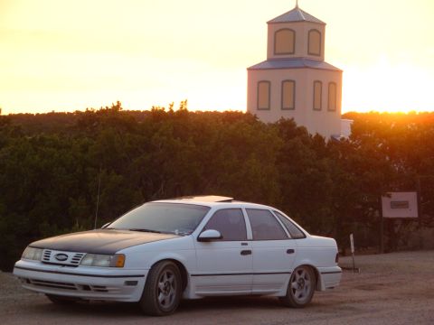 1990 Ford Taurus - A white 1990 Ford Taurus with a black hood sits parked at sunset near a distinctive tower.