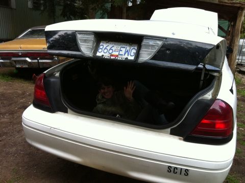 2005 Ford Crown Victoria - A person waves from inside the open trunk of a white 2005 Ford Crown Victoria, with another car visible in the background.
