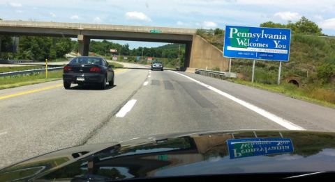 1999 Ford Crown Victoria - A black sedan travels down a highway, passing a 'Pennsylvania Welcomes You' sign.