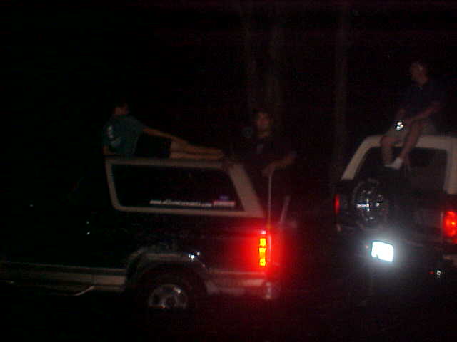 1996 Ford Bronco side view, dark exterior, with a person lying on the roof and another sitting on the roof of a second SUV