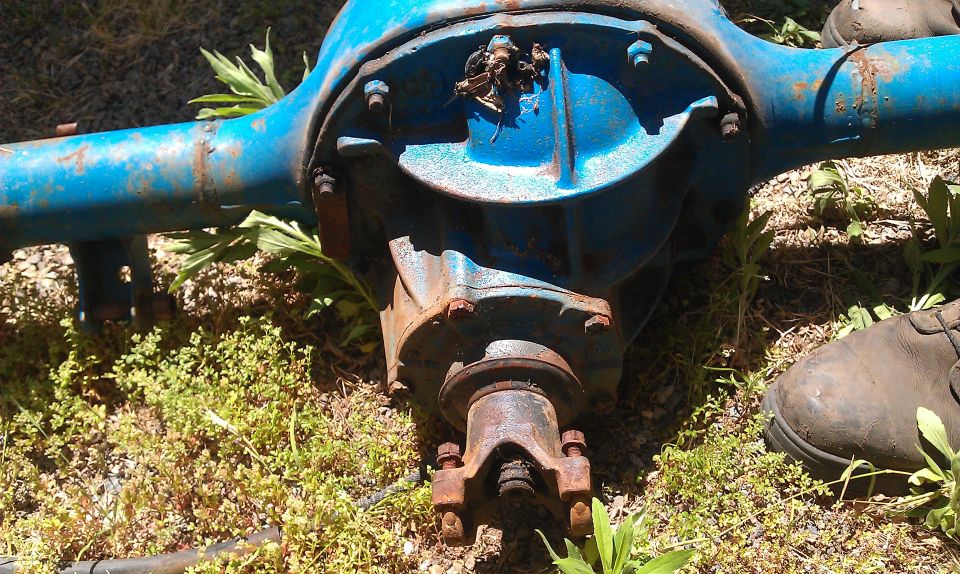 Blue painted rear axle differential assembly with U-joint yoke, resting on grass. A work boot is visible on the right