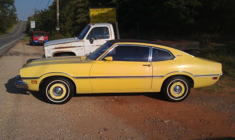 1965 Ford Mustang - A yellow 1973 Ford Maverick coupe is parked next to a white truck.