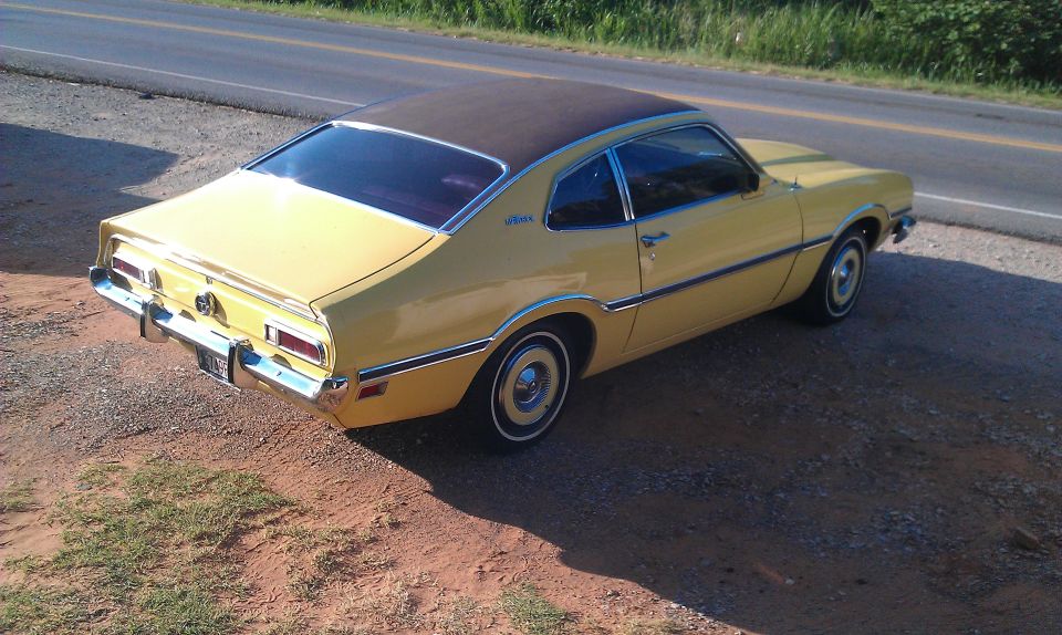 Yellow coupe with a brown roof parked on a gravel shoulder next to a road. It has a chrome bumper and trim