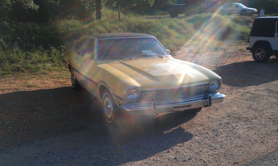 Yellow 1970 Ford Maverick with a black vinyl roof, parked on a gravel lot