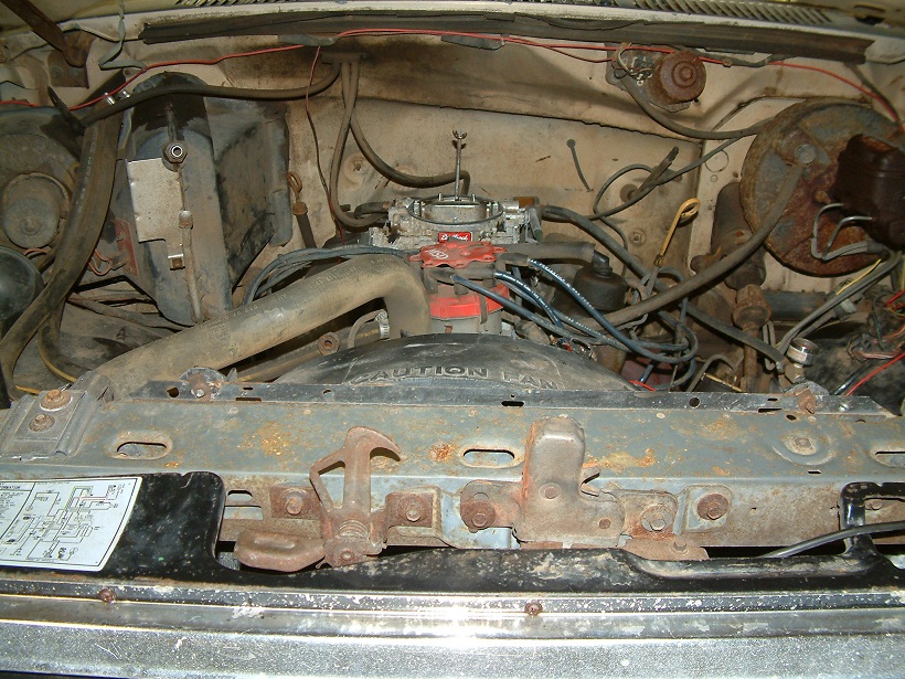 Engine bay with carburetor, hoses, and wiring. Rusty hood latches visible