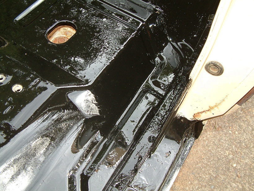 Black undercoating on vehicle floor pan with visible rust and a circular opening