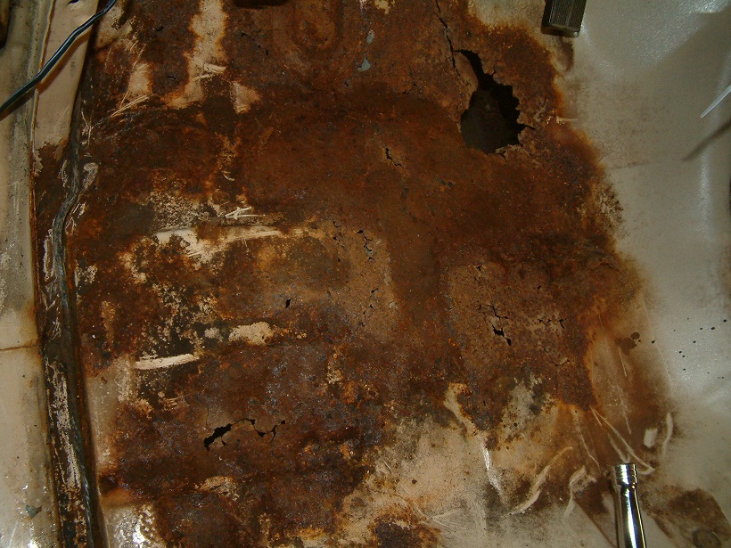 Rusted floor pan with holes, showing extensive corrosion and some white undercoating