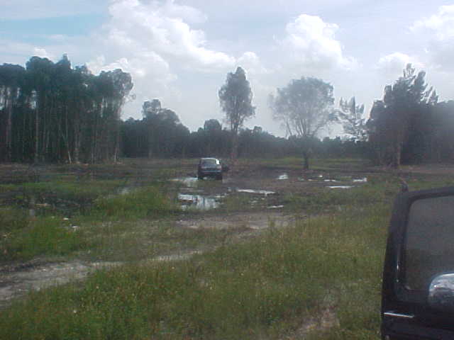 Off-road vehicle parked in a muddy field with trees in the background. Another vehicle is partially visible in the foreground