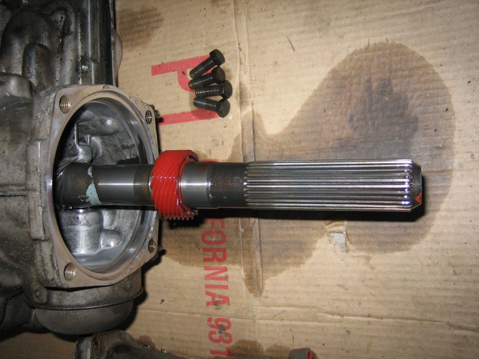 Driveshaft with splines and a red gear, next to four bolts on a cardboard surface