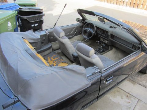 1985 Ford Mustang - The interior of a black convertible shows worn seats and a torn rear cushion, with trash bins in the background.