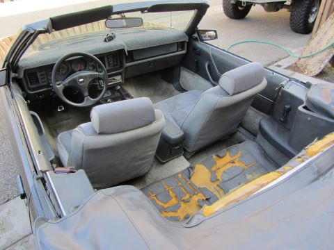 1985 Ford Mustang - The interior of a convertible shows worn upholstery and a dashboard with instrumentation. A truck is parked nearby.