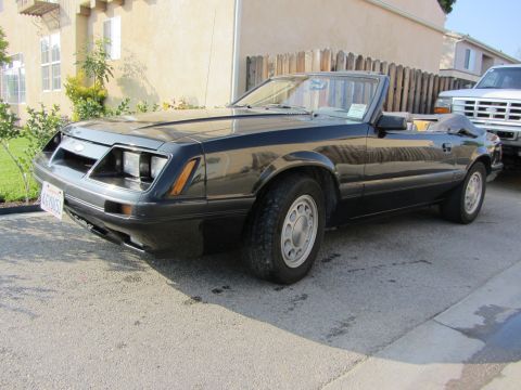 1985 Ford Mustang - Driver's-side front three-quarter view of a black Fox-body Ford Mustang LX convertible with the top folded down and a for-sale sign taped i…