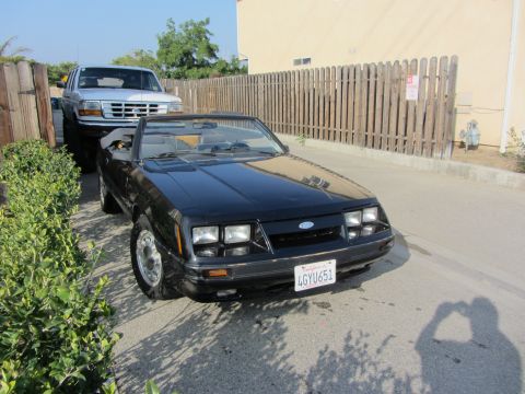 1985 Ford Mustang - High front three-quarter view of a black Fox-body Ford Mustang GT convertible with the top down, parked on a driveway beside a wood fence a…