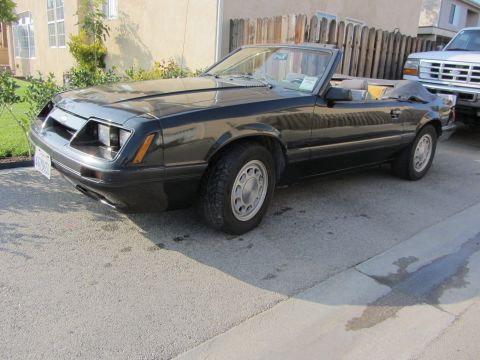 1985 Ford Mustang - Black Fox-body Ford Mustang LX convertible with the top down and a white paper sign in the windshield, shot from the driver's-side front th…