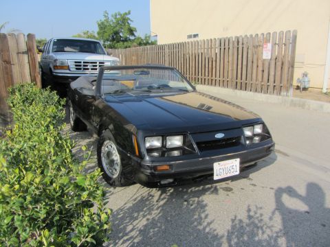 1985 Ford Mustang - Black Fox-body Ford Mustang GT convertible with the top folded down, photographed from the front passenger three-quarter angle on a drivewa…