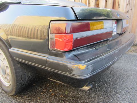 1985 Ford Mustang - Close-up of the passenger-side rear corner of a black Fox-body Ford Mustang, showing the three-segment tail light with a cracked lens patch…