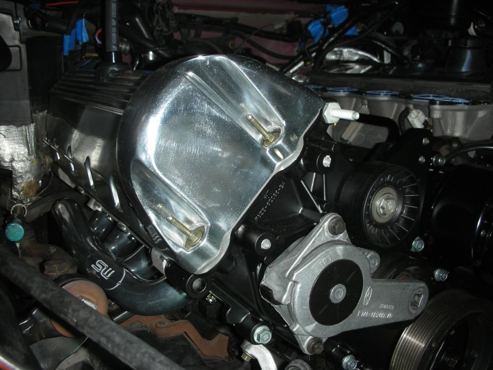 Close-up of a car engine showing a polished aluminum intake manifold bolted to the valve cover. A serpentine belt and pulley system is visible