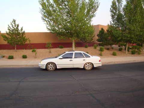 1990 Ford Taurus - A white sedan with a contrasting hood sits parked on the side of a road with trees and a building in the background.