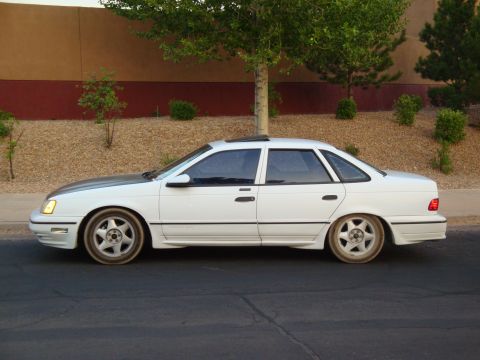 1990 Ford Taurus - A white 1990 Ford Taurus sedan with a dark hood and aftermarket wheels is seen from the side.