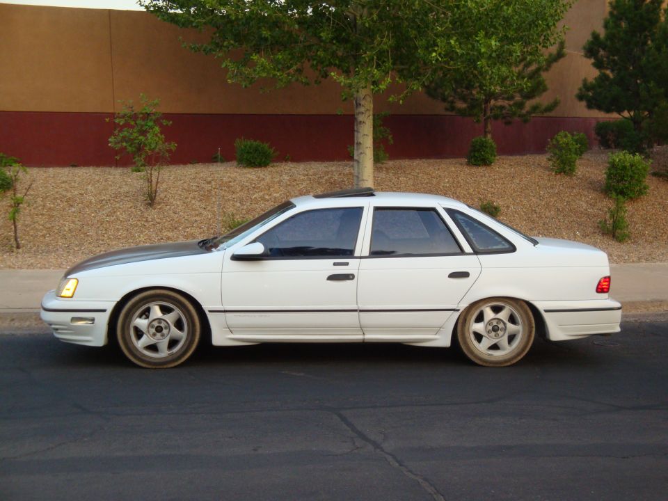 White 1990 Ford Taurus sedan with a dark hood and aftermarket wheels parked on a street