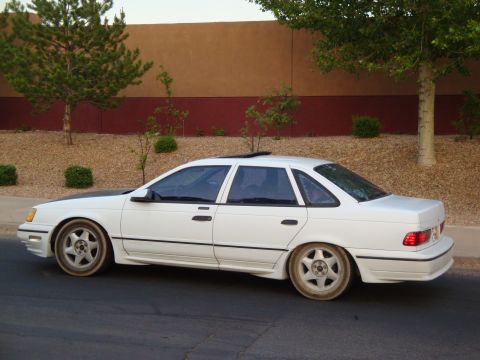 1990 Ford Taurus - A white sedan is parked on the side of the road with a textured wall and trees in the background.