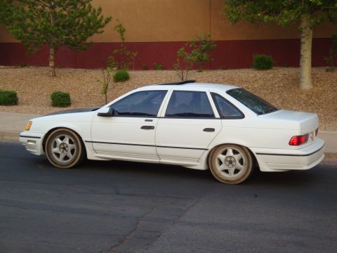 1990 Ford Taurus - A white sedan with a black hood is parked on the side of the road.