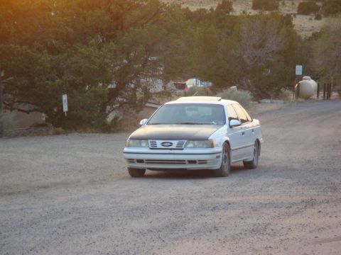 1990 Ford Taurus - White 1990 Ford Taurus sedan with a black hood travels on a dusty road in a rural landscape.
